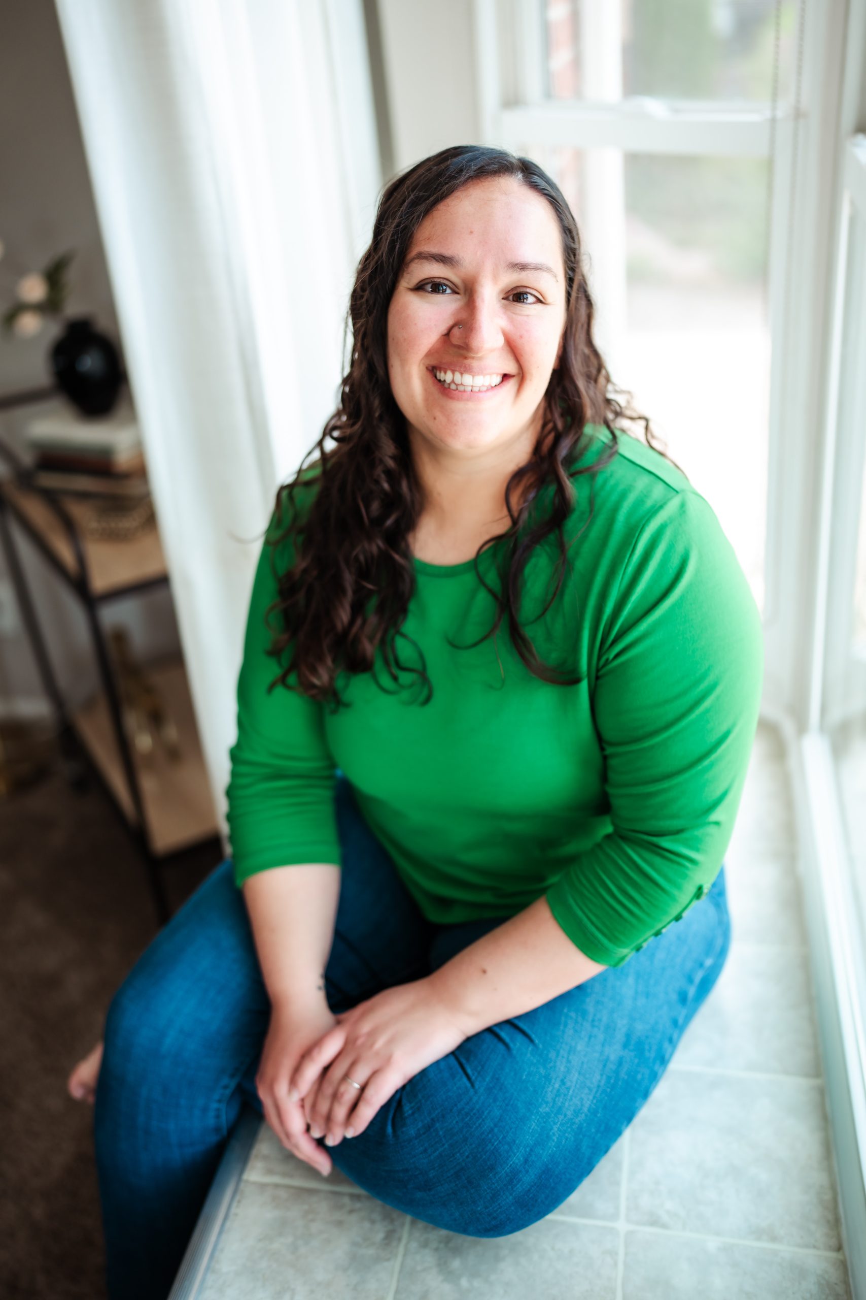 Kayla McIntosh, Birth Doula, Yoga Instructor, and owner of Whole Spirit Riverdale with wavy brown hair, wearing a rust-colored sweater and jeans, stands smiling looking over her shoulder beside a closed fence during golden hour. Reindeer are visible in the background. Connect with Kayla!